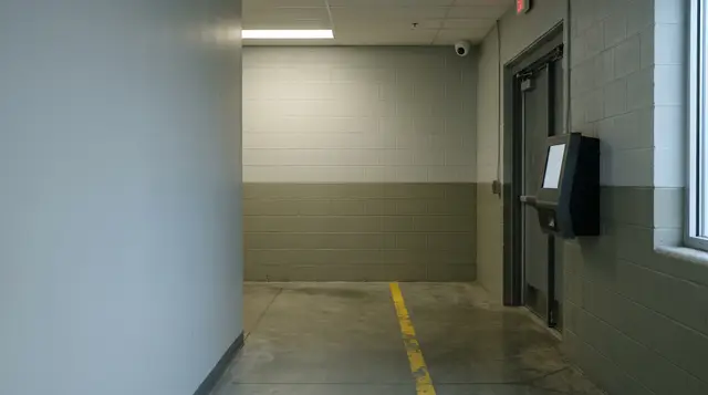 An empty industrial employee entrance corridor with a blank wall-mounted time clock beside a steel door and a small dome camera in the ceiling corner.