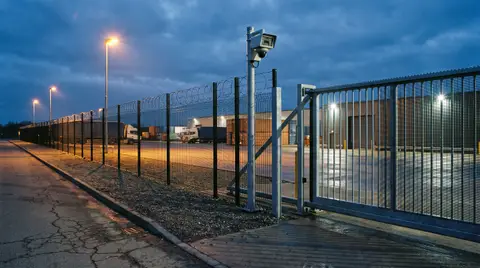 An industrial commercial facility perimeter fence line and automated gate at dusk with mounted security cameras and crisp exterior lighting.