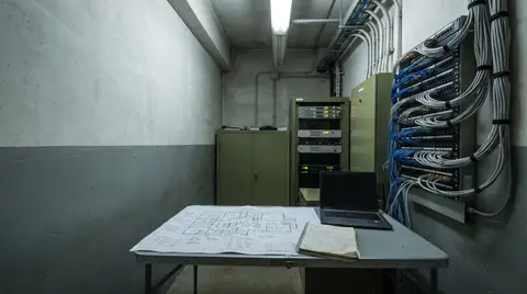 A network equipment room in an industrial facility, with a wall-mounted patch panel of dressed ethernet cabling and an annotated printed floor plan open on a table in the foreground.